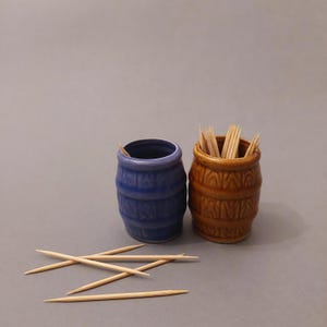 May include: Two ceramic toothpick holders shaped like barrels. One is blue and the other is brown. Toothpicks are in the brown barrel.