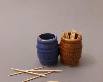 May include: Two ceramic toothpick holders shaped like barrels. One is blue and the other is brown. Toothpicks are in the brown barrel.