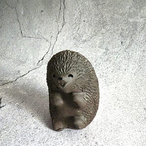 Puede incluir: Pequeña figura de erizo de cerámica marrón con una cara sonriente. El erizo tiene una espalda texturizada y puntiaguda y patas pequeñas y redondeadas. La figura está de pie y parece hecha a mano. La estética general es rústica y encantadora.