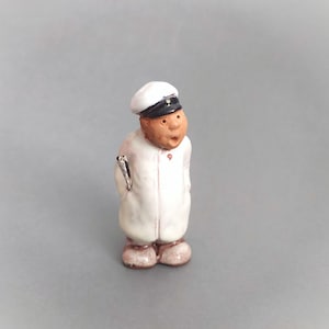 May include: A ceramic figurine of a person wearing a white uniform and a black cap. The figurine has a surprised expression and is holding a small object in its hand.