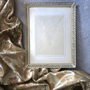 May include: A rectangular gold-colored picture frame with an ornate, openwork design around the edges. The frame holds a cream-colored mat and a blank, off-white paper. The frame rests on a gold and cream-colored patterned fabric.