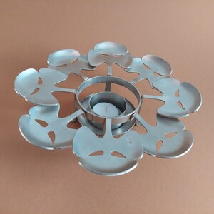 May include: Silver metal tea light candle holder with a circular base and six leaf-shaped arms. The holder is designed to hold a tea light candle in the center.