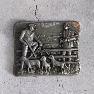 Può includere: Una fibbia per cintura in metallo argentato che raffigura due cacciatori con fucili e tre cani in un ambiente boschivo.