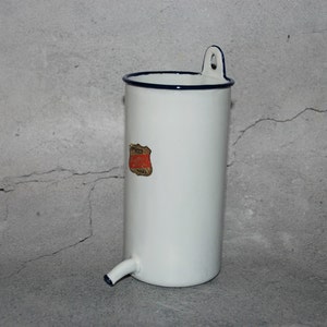Può includere: Contenitore in smalto bianco con bordo e beccuccio blu scuro. Una piccola etichetta rossa e dorata è apposta sul lato. Il contenitore ha un manico e un beccuccio. Il design generale è cilindrico.