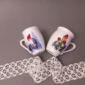 May include: Two white ceramic mugs with handles, featuring illustrations of gnomes in winter scenes. One mug shows a gnome with a black dog, while the other shows two gnomes with a red tulip.