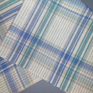May include: A close-up of a fabric with a plaid pattern in shades of blue, green, and beige. The fabric has a woven texture and a subtle sheen.