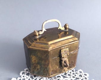 Può includere: Una scatola vintage in ottone con coperchio incernierato e manico. La scatola ha una forma ottagonale e una chiusura a fermaglio. Anche il manico e gli elementi decorativi sono in ottone. La scatola è appoggiata su un centrino di pizzo bianco.