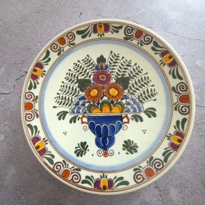 May include: A ceramic plate with a hand-painted floral design. The central image is a blue vase filled with orange, red, and blue flowers, surrounded by green leaves. The rim has a blue border and a repeating floral pattern.