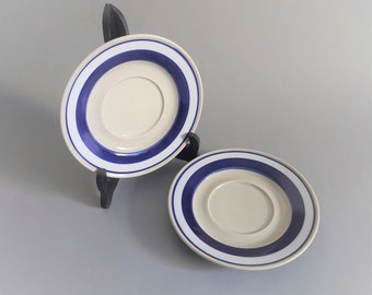 May include: Two white ceramic saucers with blue stripes. The saucers are stacked on top of each other.