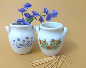 May include: Two small, white ceramic vases with handles, one with blue floral designs and the other with orange and green berry patterns. Blue flowers are arranged in one vase. Wooden toothpicks are scattered in front of the vases.
