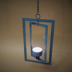 May include: A hanging candle holder with a white metal frame. The frame is made of two squares, one inside the other, creating a geometric design. A small white candle sits inside the inner square.