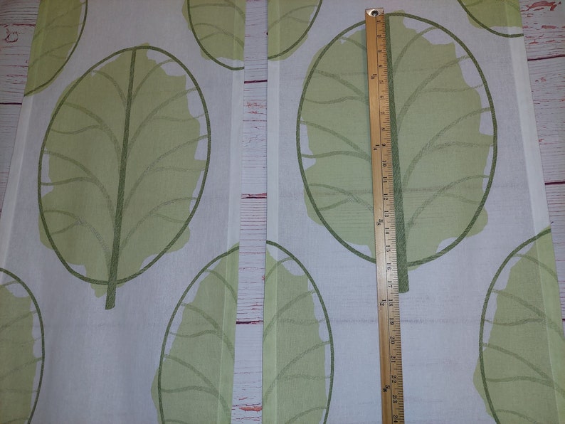 May include: Two panels of fabric with a repeating pattern of large, light green leaves on a white background. A wooden ruler is placed vertically, showing measurements up to 24 inches.