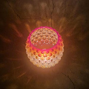 May include: A spherical, decorative candle holder with a textured, bubble-like surface. The color gradient transitions from white at the base to orange and pink at the top. The candle holder is lit from within, casting a warm glow.
