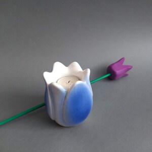 May include: A blue and white ceramic tulip candle holder with a tealight candle inside. The holder is sitting on a gray surface with a purple tulip stem.