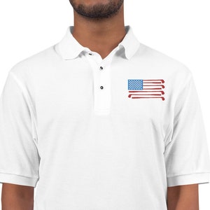 Puede incluir: Camiseta polo blanca con un dise&ntilde;o de bandera americana roja, blanca y azul hecha de palos de golf. La bandera est&aacute; en el pecho izquierdo de la camisa.