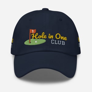 May include: A navy blue baseball cap with an embroidered green golf course, a red flag with a white "1" on it, a white golf ball, and the text "Hole in One Club" in yellow.