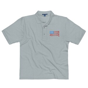 Puede incluir: Una camiseta polo gris claro con un dise&ntilde;o de bandera estadounidense roja, blanca y azul hecha de palos de golf en el pecho izquierdo.