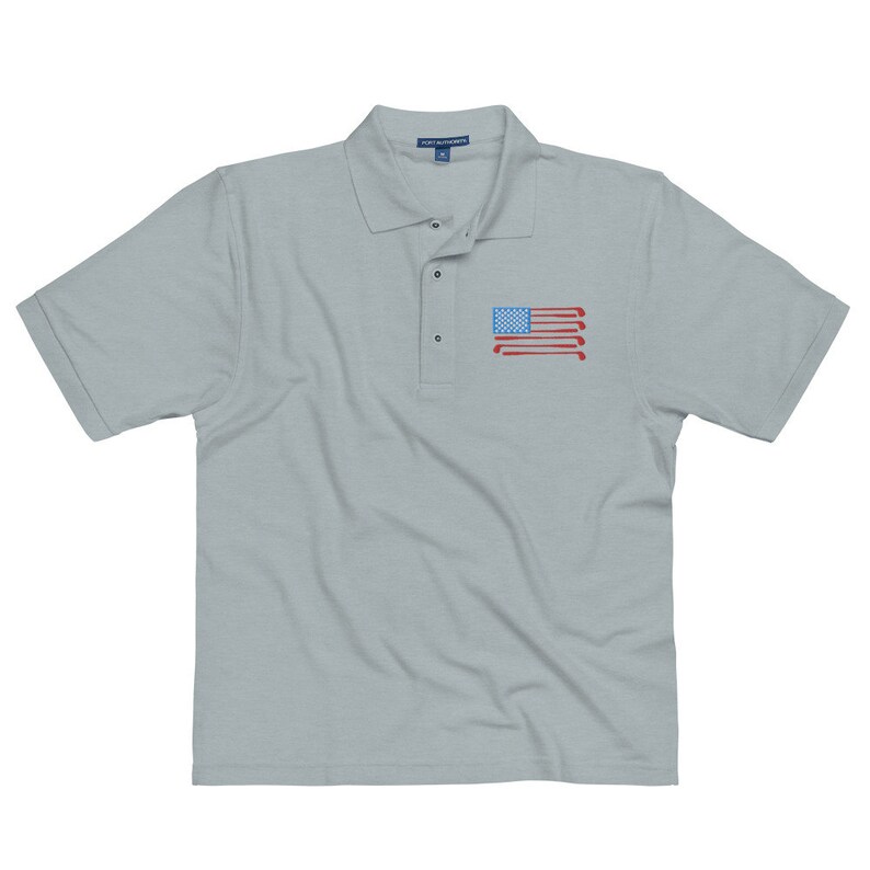 Puede incluir: Una camiseta polo gris claro con un dise&ntilde;o de bandera estadounidense roja, blanca y azul hecha de palos de golf en el pecho izquierdo.