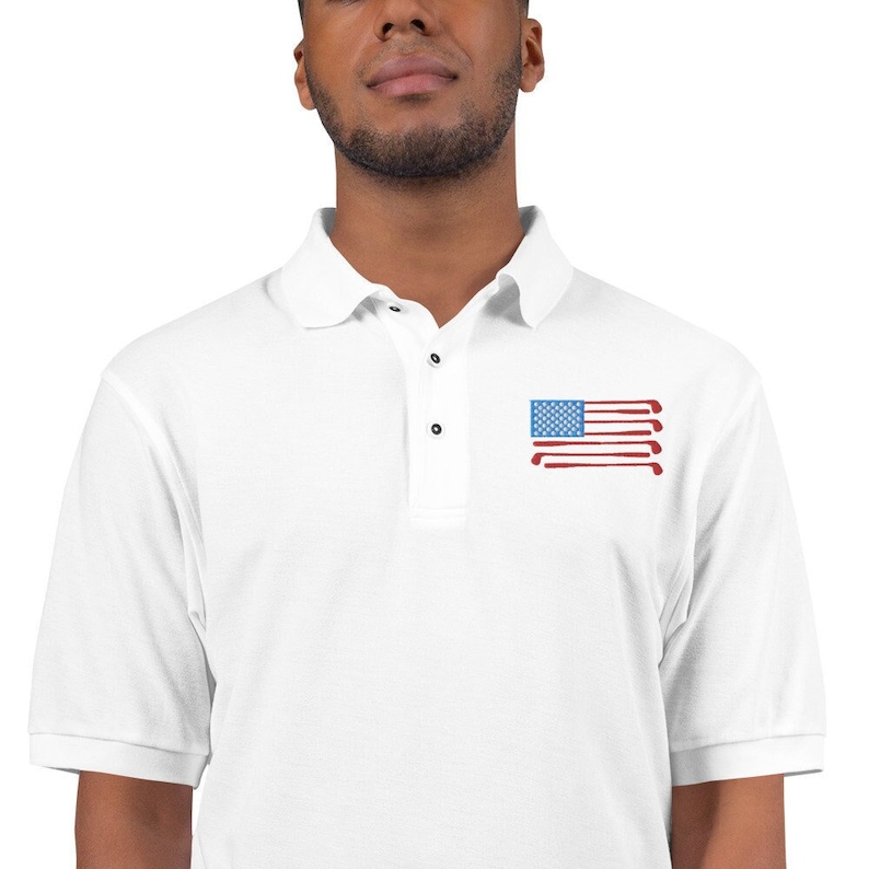 Puede incluir: Camiseta polo blanca con un dise&ntilde;o de bandera americana roja, blanca y azul hecha de palos de golf. La bandera est&aacute; en el pecho izquierdo de la camisa.