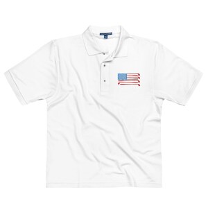 Puede incluir: Polo blanco con un dise&ntilde;o de bandera estadounidense roja, blanca y azul hecho de palos de golf en el pecho izquierdo.