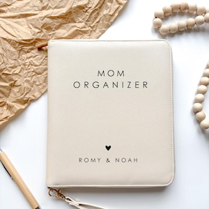Könnte beinhalten: Cremefarbener Mom Organizer mit Reißverschluss und Handschlaufe. Der Text "MOM ORGANIZER" ist in Schwarz gedruckt, zusammen mit einem kleinen Herzen und den Namen "ROMY & NOAH". Ein Holzstift und eine Perlenkette befinden sich im Hintergrund.
