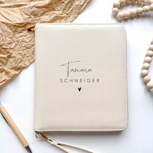 Könnte beinhalten: Cremefarbenes Portfolio mit Reißverschluss, auf dem der Name "Tamara Schneider" und ein kleines Herz in Schwarz aufgedruckt sind. Das Portfolio hat eine Handschlaufe und goldfarbene Hardware. Ein Holzstift und eine Kette aus Holzperlen sind ebenfalls zu sehen.
