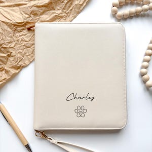 Könnte beinhalten: Cremefarbene Reißverschluss-Clutch mit dem Namen "Charley" und einem Pfotenabdruck-Design. Die Clutch hat eine Handschlaufe und goldfarbene Hardware. Ein Holzstift und eine Kette aus Holzperlen sind ebenfalls zu sehen.