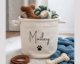 May include: A cream-colored canvas storage basket with rope handles, featuring the name Mailey and a black paw print. The basket is filled with various dog toys, including a brown plush bone, while a brown leather leash lies nearby.