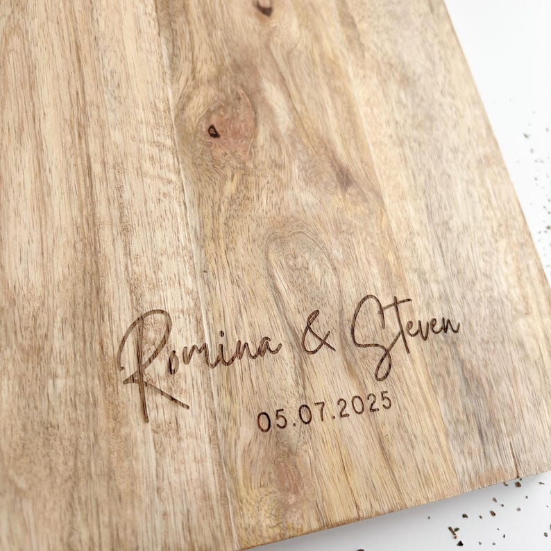 May include: A light-colored wooden cutting board with the names "Romina & Steven" and the date "05.07.2025" engraved in a cursive font. The wood grain is visible, with natural knots and patterns.