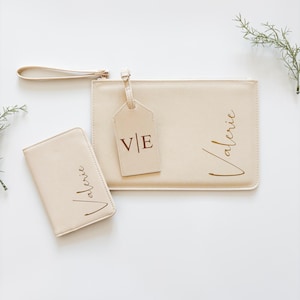 Könnte beinhalten: Ein cremefarbenes Set aus Clutch, Reisepasshülle und Gepäckanhänger. Die Clutch und die Reisepasshülle sind mit dem Namen "Valerie" in goldener Schrift versehen. Der Gepäckanhänger zeigt die Initialen "V|E". Die Clutch hat eine Handschlaufe.