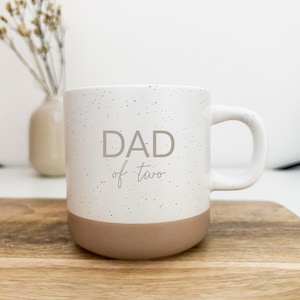 Könnte beinhalten: Weiße Keramiktasse mit gesprenkeltem Design und beigefarbenem Boden. Die Tasse hat einen gebogenen Henkel und die Worte "DAD of two" in Grau aufgedruckt. Die Tasse steht auf einer Holzoberfläche.