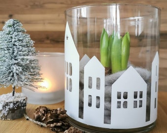 Puede incluir: Un jarrón cilíndrico de cristal transparente decorado con siluetas de casas en vinilo blanco. Dentro del jarrón, bulbos de jacinto verdes descansan sobre una base de lana gris. El jarrón está rodeado de piñas, una vela y un árbol.