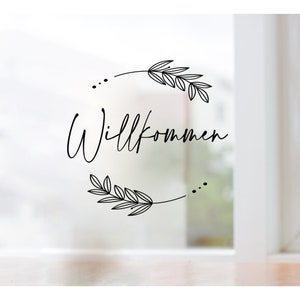 Könnte beinhalten: Ein schwarz-weißes Willkommensschild mit einem Kranz aus Blättern und dem Wort "Willkommen" in einer kursivschrift.