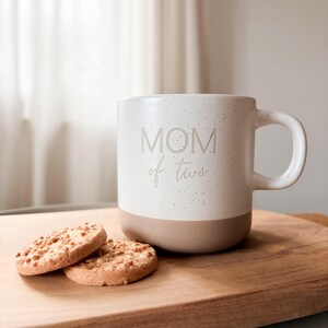 Könnte beinhalten: Eine weiße Keramiktasse mit den Worten "MOM of two" auf der Seite. Die Tasse hat ein gesprenkeltes Design und einen beigefarbenen Boden. Zwei runde Kekse mit Nüssen liegen neben der Tasse auf einem Holzschneidebrett.
