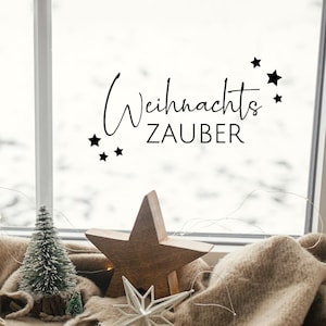 Könnte beinhalten: Ein Fenster mit einer verschneiten Szene draußen. Das Fenster hat einen dekorativen Aufkleber mit der Aufschrift "Weihnachtszauber" mit Sternen. Ein Holzstern-Anhänger und ein weißer Stern-Anhänger liegen auf einer braunen Decke unter dem Fenster.