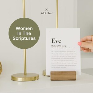 May include: A minimalist print featuring the name "Eve" and text from the scriptures, held in a wooden block stand. The image is part of a series titled "Women In The Scriptures" and includes a gold-toned lamp.