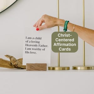 May include: A white affirmation card with the text "I am a child of a loving Heavenly Father. I am worthy of His love." is displayed on a wooden stand. A green card with "Christ-Centered Affirmation Cards" is also visible.