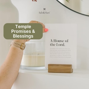 May include: A white card with the text "A House of the Lord" sits in a wooden holder. The card is held by a person with a beige watch and pink nail polish. A candle in a glass jar and a white vase are in the background.
