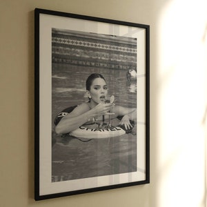 May include: A black and white photo of a person in a swimming pool. The person is wearing a swimsuit and is holding a glass of champagne. The person is floating on a pool float that says 'CHANEL'.