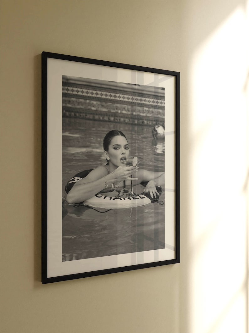 May include: A black and white photo of a person in a swimming pool. The person is wearing a swimsuit and is holding a glass of champagne. The person is floating on a pool float that says 'CHANEL'.