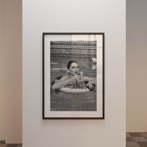 May include: A black and white photograph of a woman in a swimming pool. The woman is wearing a swimsuit and is holding a glass of champagne. The woman is floating on a white inflatable raft with the word 'CHANEL' printed on it.