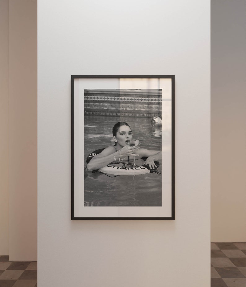 May include: A black and white photograph of a woman in a swimming pool. The woman is wearing a swimsuit and is holding a glass of champagne. The woman is floating on a white inflatable raft with the word 'CHANEL' printed on it.