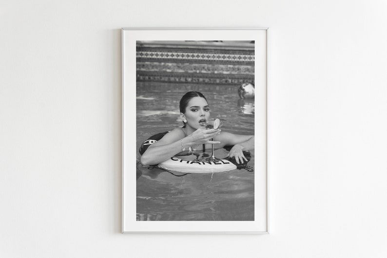 May include: A black and white photo of a woman in a pool wearing a Chanel swimsuit and a Chanel float. The woman is holding a glass of champagne and looking at the camera.