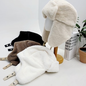 May include: A display of faux fur trapper hats in cream, black, brown, and white. The hats have ear flaps and adjustable chin straps with metal snaps. One hat is displayed on a mannequin head. The hats are designed for cold weather.