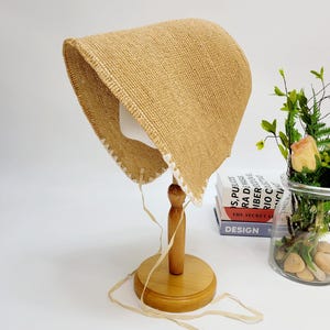 May include: A straw-colored bonnet with a white lace trim, displayed on a wooden stand. The bonnet has a rounded crown and ties with long, cream-colored ribbons. The stand is made of light-colored wood.