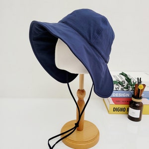 May include: A navy blue sun hat with a wide brim and a black drawstring. The hat is displayed on a wooden stand.