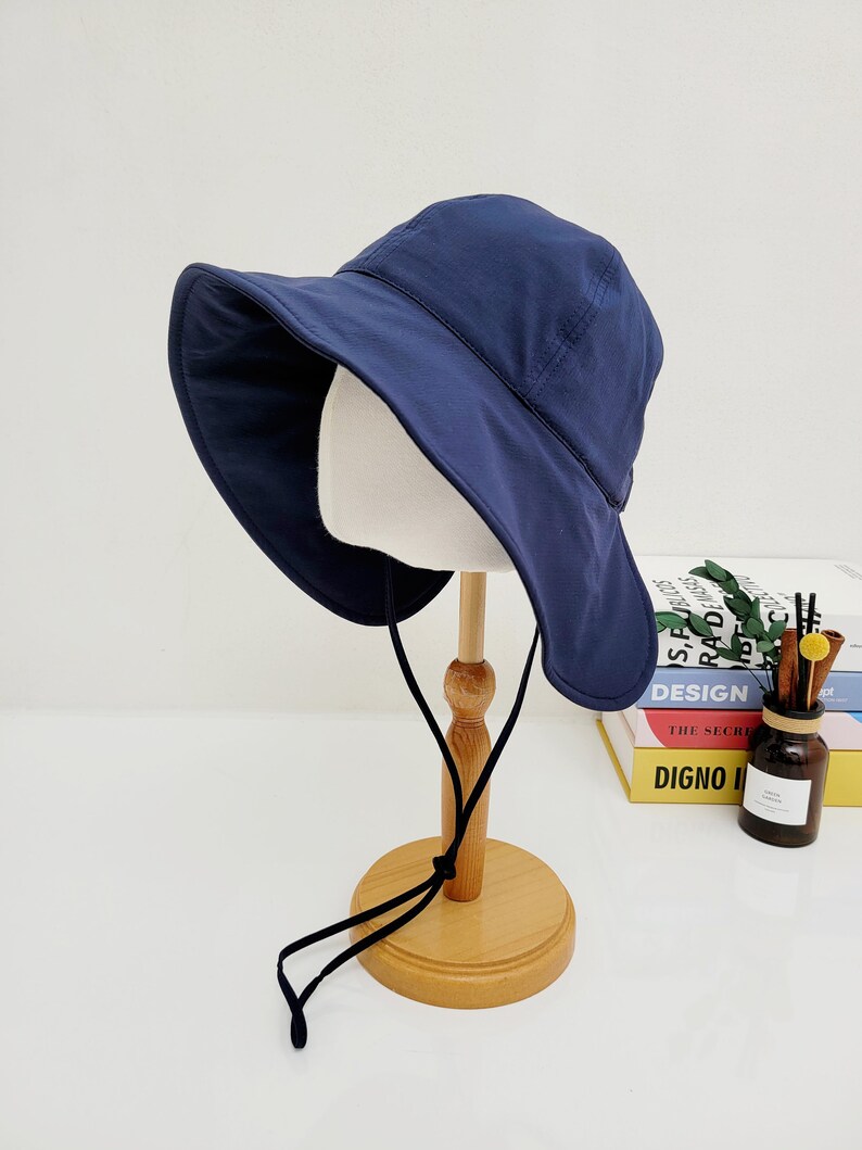 May include: A navy blue sun hat with a wide brim and a black drawstring. The hat is displayed on a wooden stand.