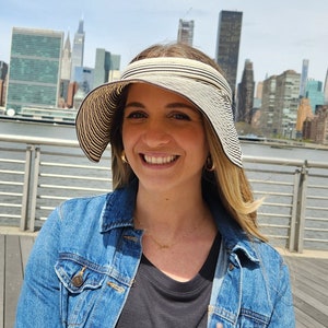 May include: A woman wearing a black and white striped sun visor with a denim jacket and a black shirt. The woman is standing in front of a city skyline.