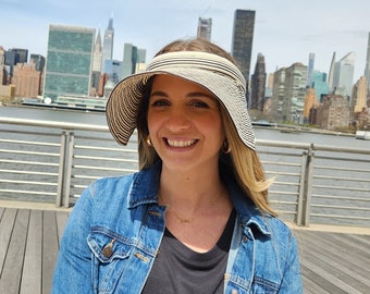 May include: A woman wearing a black and white striped sun visor with a denim jacket and a black shirt. The woman is standing in front of a city skyline.