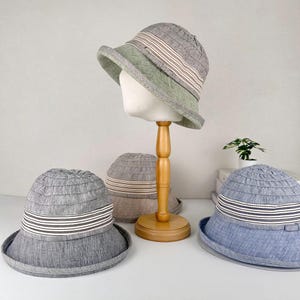 May include: A collection of stylish bucket hats in various colors, including gray, green, and blue. Each hat features a striped band and a structured brim. One hat is displayed on a mannequin head, showcasing the hat's shape and design.
